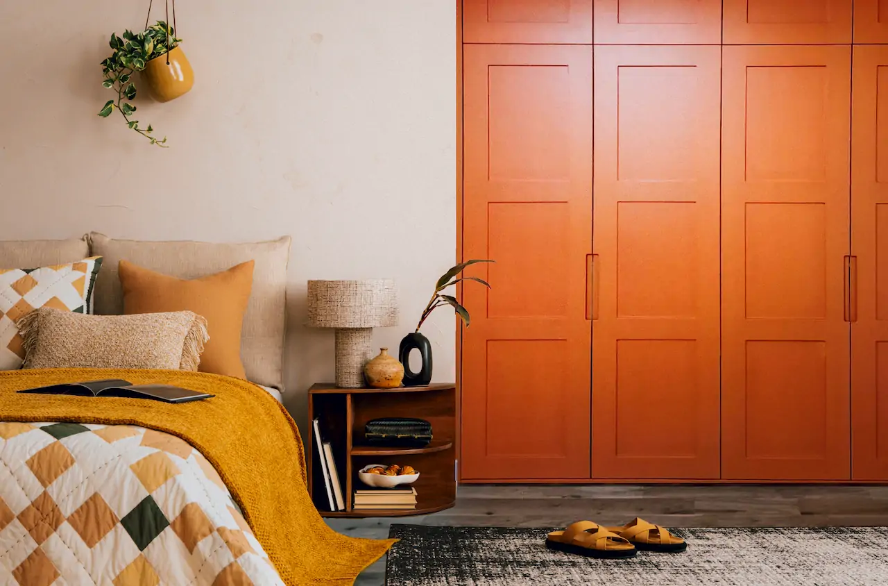 A light brown room with an orange fitted wardrobe next to a bed with orange and white covers, there are also a pair of orange slippers on a rug