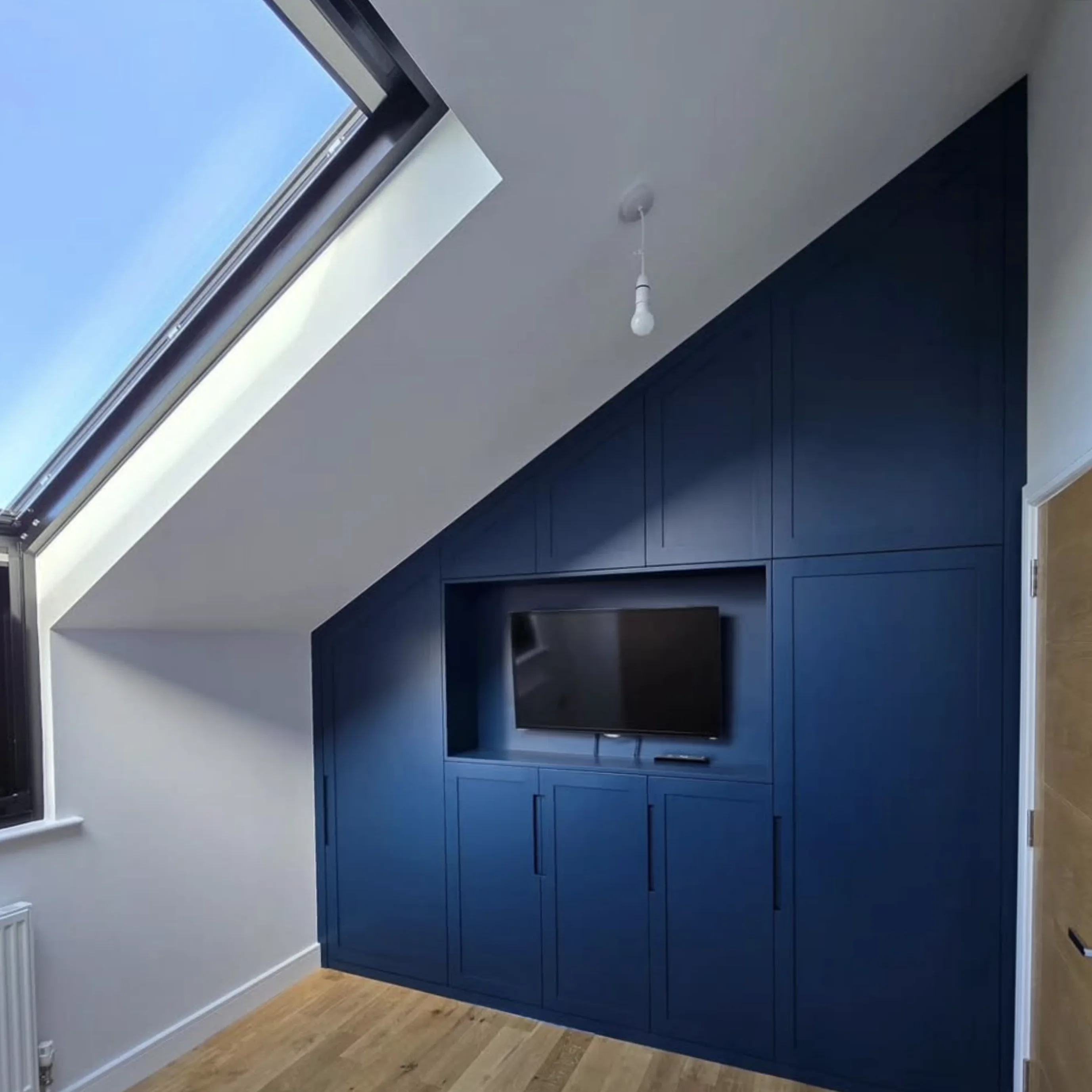 A loft bedroom with fitted blue wardrobes and built-in TV niche under a skylight
