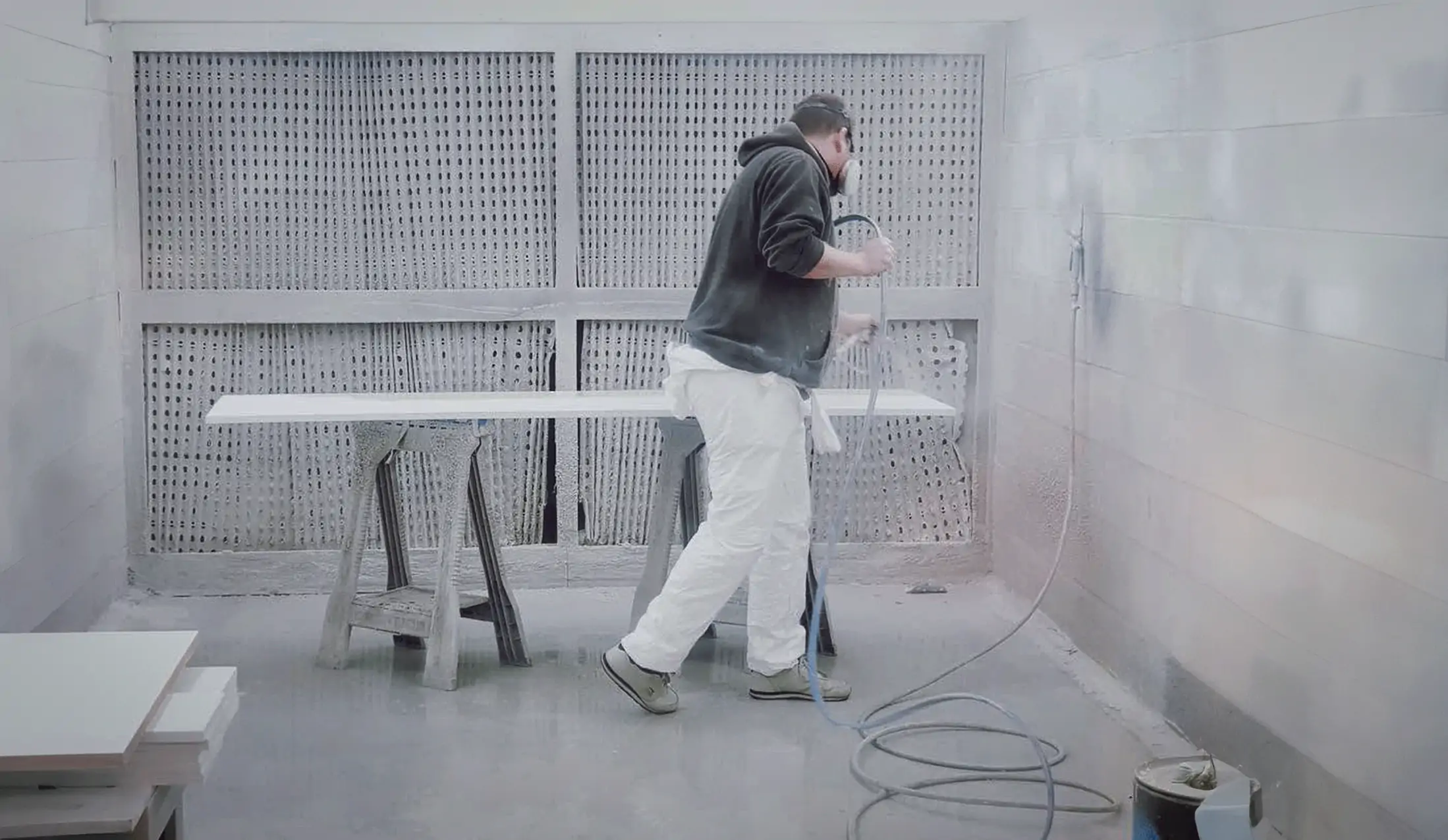 A man in white overalls sprays white paint on wood in a spray room.